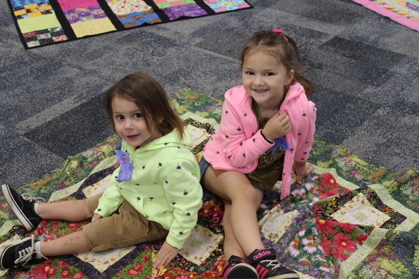 two preschool-aged girls with dark hair wearing brightly colored sweatshirts, one lime green and the other pink, sitting on a quilt and looking at the camera