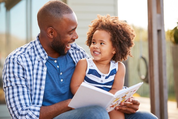 African American father wearing blue checked shirt reading a book and laughing with his toddler