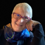 woman with short gray hair and red glasses leaning her head on her hand and smiling at camera