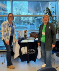 Two women with blonde hair standing in front of a table that has a hedberg public library tablecloth in front of large windows