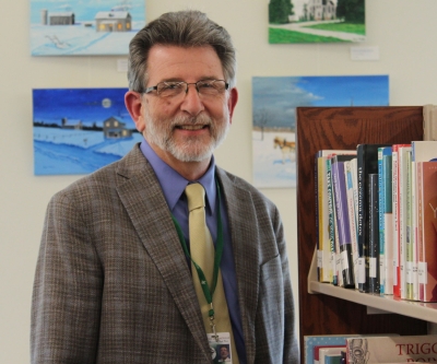 Man glasses and grey hair wearing a suit standing next to a shelf of books, smiling
