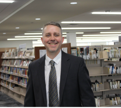 man with beard wearing suit smiling in front of book stacks