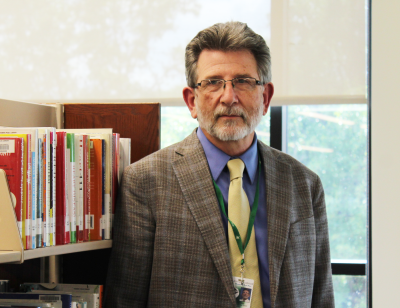 man in suit with glasses looking at camera and standing next to shelf of books