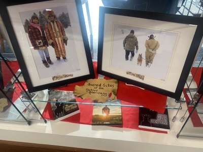 Two photos with two Native American men in each photo.  Photo on the left has two Native American men wearing traditional winter coats.  Photo on the right has two Native American men at the Snow Snake games on Madeline Island with a dog walking away from the camera.   Books about sacred Native American sites on a glass shelf below the photos.