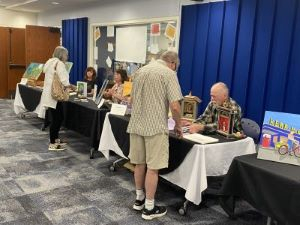 Three adults sitting behind long tables topped with their artwork. Two adults in forefront chat with artists. 