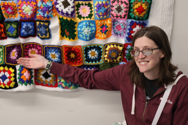 Woman with dark hair and glasses smiling at camera and pointing to crocheted blanket