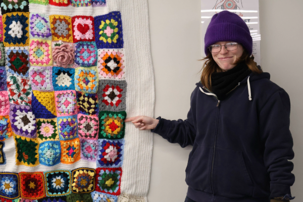 woman wearing purple hat and glasses smiling at the camera and pointing to a crocheted blanket