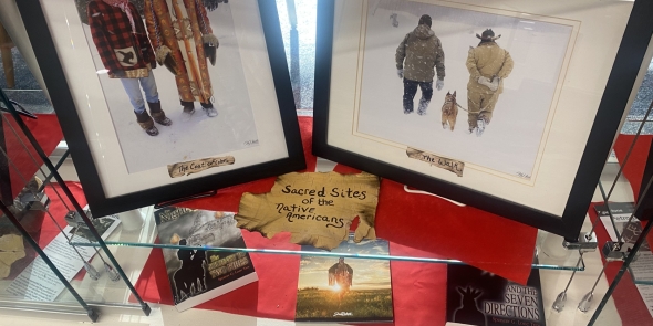 Two photos with two Native American men in each photo.  Photo on the left has two Native American men wearing traditional winter coats.  Photo on the right has two Native American men at the Snow Snake games on Madeline Island with a dog walking away from the camera.   Books about sacred Native American sites on a glass shelf below the photos.