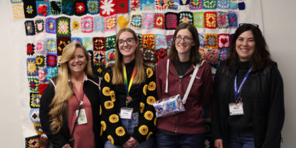 Four women smiling at the camera standing in front of a crocheted blanket hanging on the wall