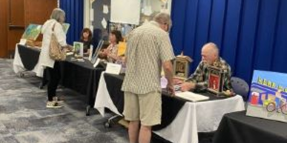 Three adults sitting behind long tables topped with their artwork. Two adults in forefront chat with artists. 
