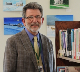 Man glasses and grey hair wearing a suit standing next to a shelf of books, smiling