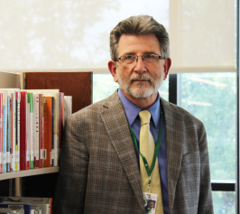 man in suit with glasses looking at camera and standing next to shelf of books