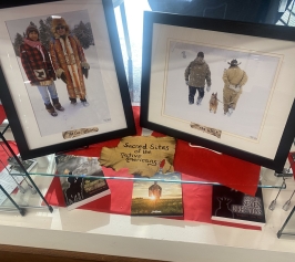 Two photos with two Native American men in each photo.  Photo on the left has two Native American men wearing traditional winter coats.  Photo on the right has two Native American men at the Snow Snake games on Madeline Island with a dog walking away from the camera.   Books about sacred Native American sites on a glass shelf below the photos.