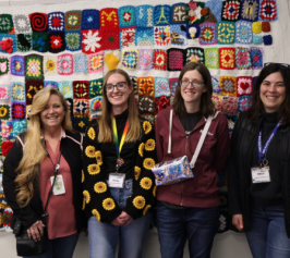 Four women smiling at the camera standing in front of a crocheted blanket hanging on the wall