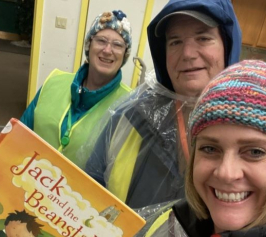 Three adults in warm rain gear. Woman in forefront holds book Jack and the Beanstalk.