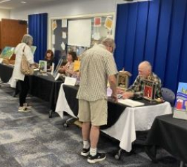 Three adults sitting behind long tables topped with their artwork. Two adults in forefront chat with artists. 