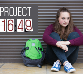Teen with backpack looking despondent, sitting on sidewalk. 
