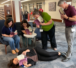 Two adults sitting reading and two adults standing in a library with author Maggie Ginsberg reading book on the floor.