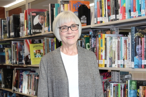 woman with gray hair and glasses wearing a gray sweater cardigan standing next to a bookshelf