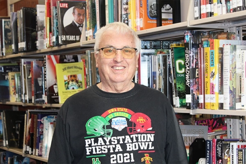 man with gray hair and glasses wearing a football tshirt standing next to a bookshelf