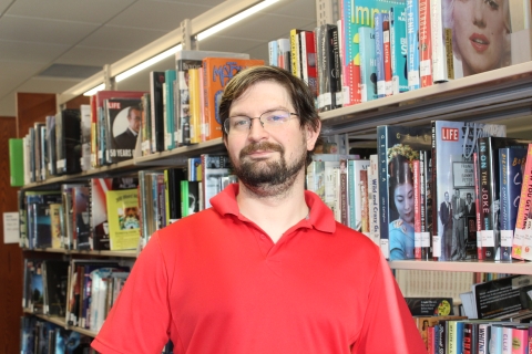 man with brown hair and beard wearing glasses and a red polo shirt standing next to a bookshelf