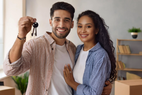 latino couple smiling at the camera holding keys and standing in front of moving boxes inside their house