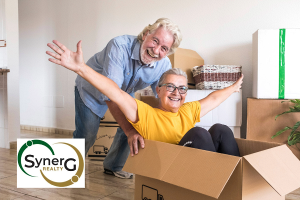 Older woman in yellow shirt in packing box with arms up in celebration. Older man behind her.