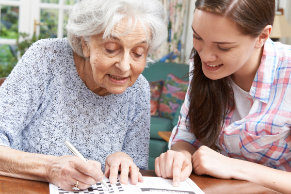 older woman with gray hair doing a crossword puzzle while a younger woman watches and smiles