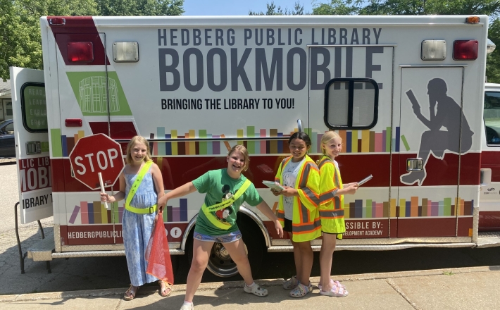 four young students standing in front of the library bookmobile holding a stop sign and wearing safety vests