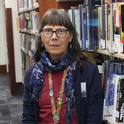 Woman with long dark hair wearing black glasses, a blue scarf, red shirt and a blue cardigan leaning on a book shelf