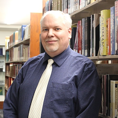 man with gray hair and goatee wearing a purple shirt and yellow tie standing next to a bookshelf