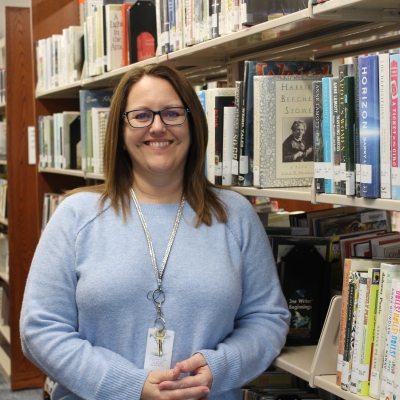 woman with brown hair and glasses wearing a light blue sweater standing next to book shelf