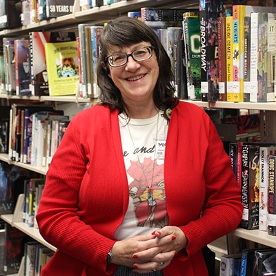 Woman with dark hair and glasses wearing a red cardigan leaning on a bookshelf