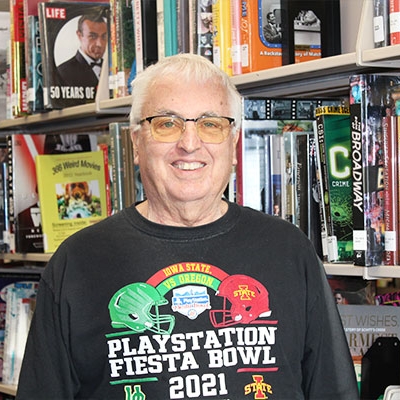 man with gray hair and glasses wearing a football tshirt standing next to a bookshelf