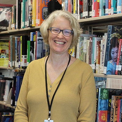 Blonde woman in glasses wearin a yellow top leaning against a bookshelf, smiling