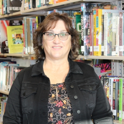 woman with dark hair and glasses wearing a black denim jacket leaning against a bookshelf