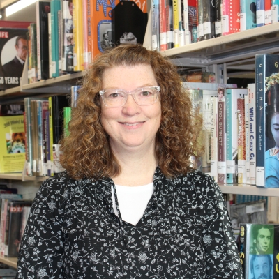 woman with brown hair and glasses wearing a black and white shirt standing next to a bookshelf