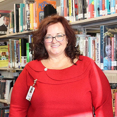woman with dark brown hair and glasses wearing a red sweater standing next to a bookshelf