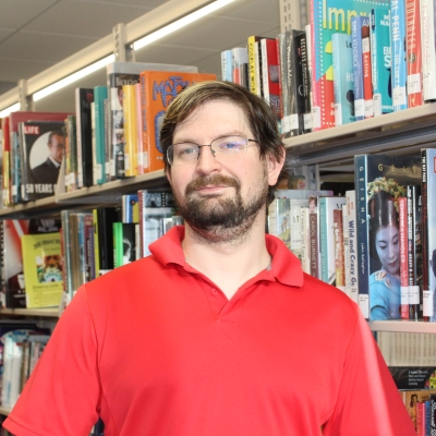 man with brown hair and beard wearing glasses and a red polo shirt standing next to a bookshelf