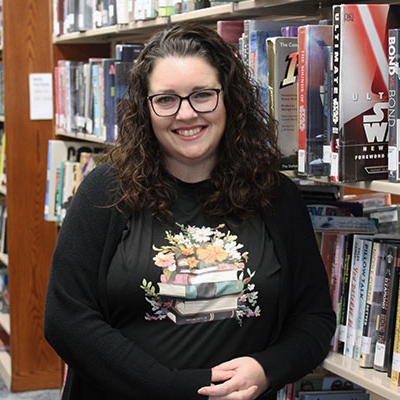 woman with dark curly hair and glasses wearing a black cardigan and black tshirt with books on it leaning on a bookshelf