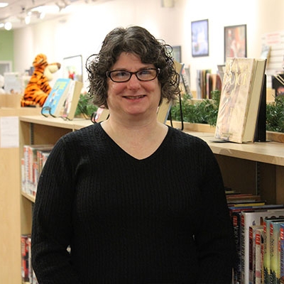 woman with short brown curly hair wearing glasses and a black shirt standing next to a book shelf