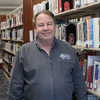 man with brown hair wearing denim button up standing next to book shelf
